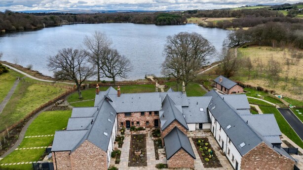 A large, multi-story brick building with a symmetrical roofline, surrounded by a well-maintained lawn and a lake in the background. - Tarn End Cottage 5, Talkin Tarn, ideal for a family, activities and historical sites nearby (BRAMPTON)