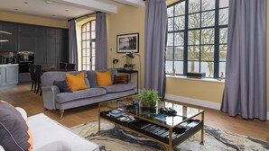 A modern living room with a grey sofa, a coffee table, and a view of a kitchen and dining area through large windows. - Tarn End Cottage 5, Talkin Tarn, ideal for a family, activities and historical sites nearby (BRAMPTON)