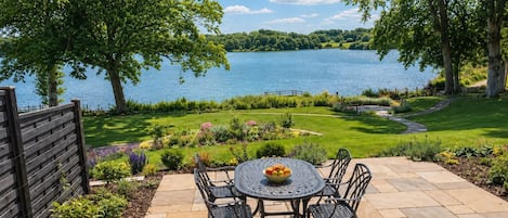 An outdoor patio with a round table and chairs, surrounded by a well-maintained garden and a view of a lake.