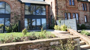 A two-story brick house with large windows, a stone retaining wall, and a small garden area. - Tarn End Cottage 8, Talkin Tarn, Brampton ideal for a romantic break overlooking the Tarn (BRAMPTON)