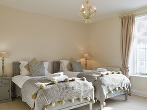 A bedroom with two beds, bedside lamps, a chandelier, and a window with curtains.