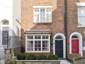 A two-story brick building with a white window and door, a red door, and a small garden area with bushes.