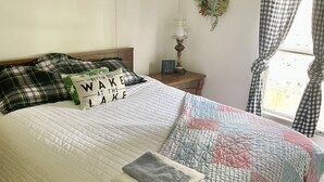 A neatly made bed with a quilt and pillows, a wall-mounted lamp, a checkered curtain, and a small plant on the wall.