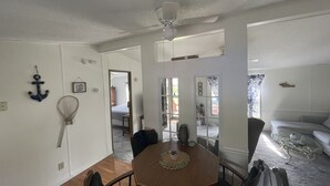 A dining area with a wooden table, chairs, and a ceiling fan. There is a view of a living room with a couch and a glass coffee table.