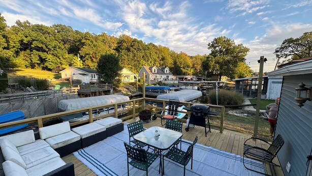 A deck with outdoor furniture, a grill, and a view of a lake and houses in the background.