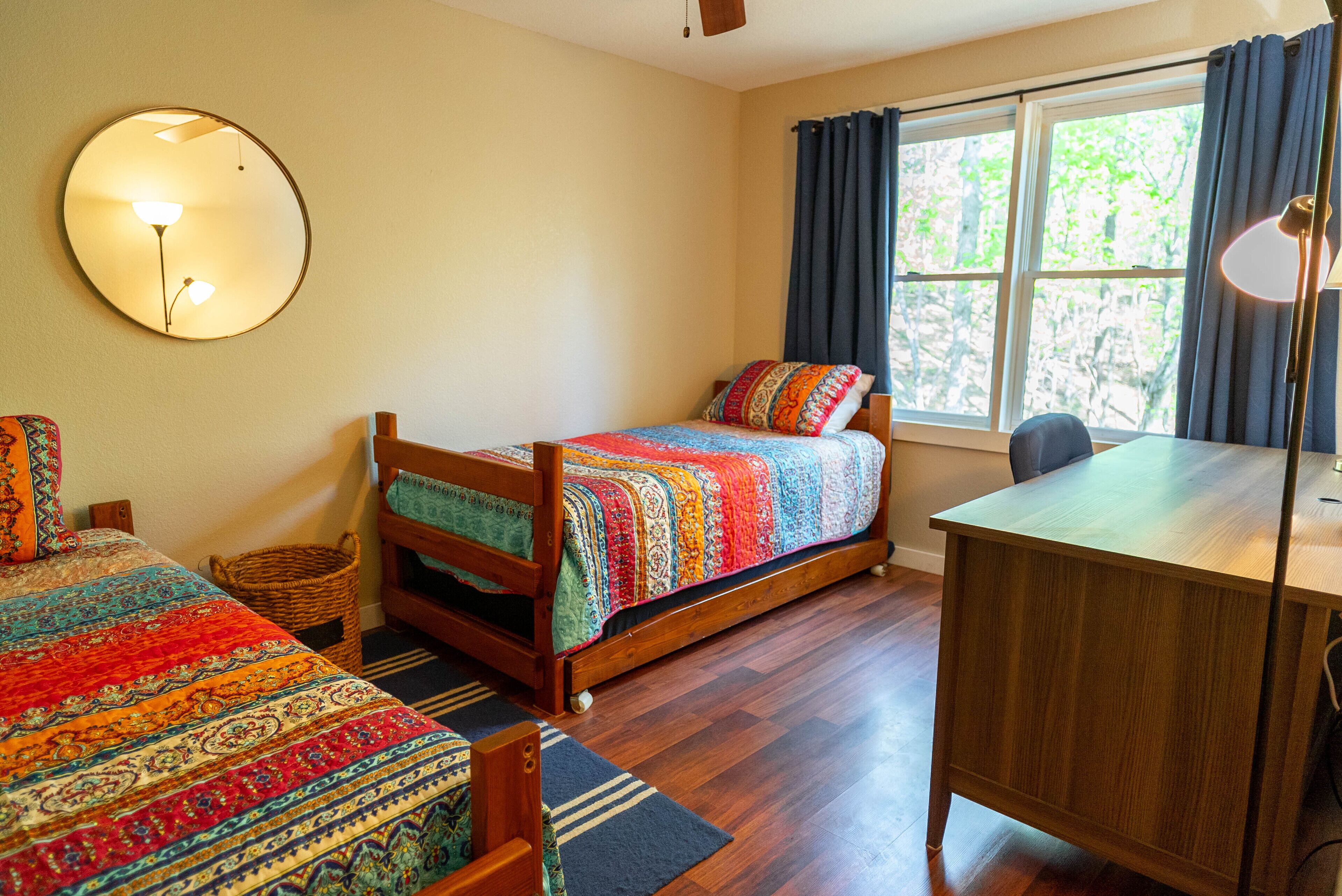 A bedroom with a bed, a desk, a chair, a mirror, and a window with curtains.