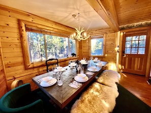 A wooden cabin dining room with a long table set for four, large windows, and a chandelier. - Enchanted Log Home on Large Lot Pet friendly (Big Bear Lake)