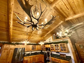A rustic cabin interior with wooden ceilings, a large antler chandelier, wooden cabinets, and a stainless steel refrigerator. - Enchanted Log Home on Large Lot Pet friendly (Big Bear Lake)