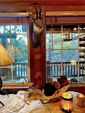 A room with a mounted animal head, a wooden shelf, a window with a view of trees, and a candle on a table. - Enchanted Log Home on Large Lot Pet friendly (Big Bear Lake)