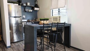 A compact kitchen with a stainless steel refrigerator, a microwave, and a gas stove. There are black chairs around the island, and a small potted plant on the counter. - The White Bungalow (Rosarito)