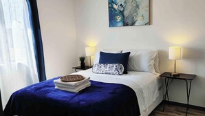 A neatly arranged bedroom with a bed, two bedside tables, a lamp, and a painting on the wall. - The White Bungalow (Rosarito)