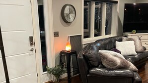 A living room with a black leather sofa, a small side table with a lamp, a clock on the wall, and a large window.