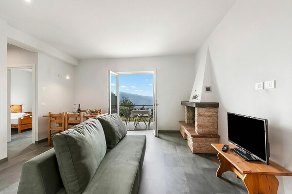 A modern living room with a sofa, a TV, and a fireplace. There is a balcony with a view of the sky and mountains.