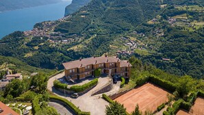 A large, multi-story building with a tennis court, surrounded by lush greenery and mountains.