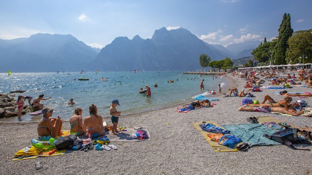 A crowded beach with people sunbathing and swimming, surrounded by mountains.