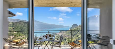 A balcony with a table set for dining, a chair, and a view of a mountainous landscape and lake.