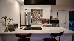 Una cocina moderna con un refrigerador de acero inoxidable, un microondas y una cafetera sobre la encimera. Hay dos sillas en la isla y una maceta colgante arriba.