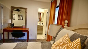 Habitación de hotel con cama, escritorio, silla, espejo y baño visible a través de una puerta abierta.