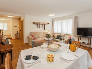 A dining area with a table set for four, a piano, a sofa, and a television in the background.
