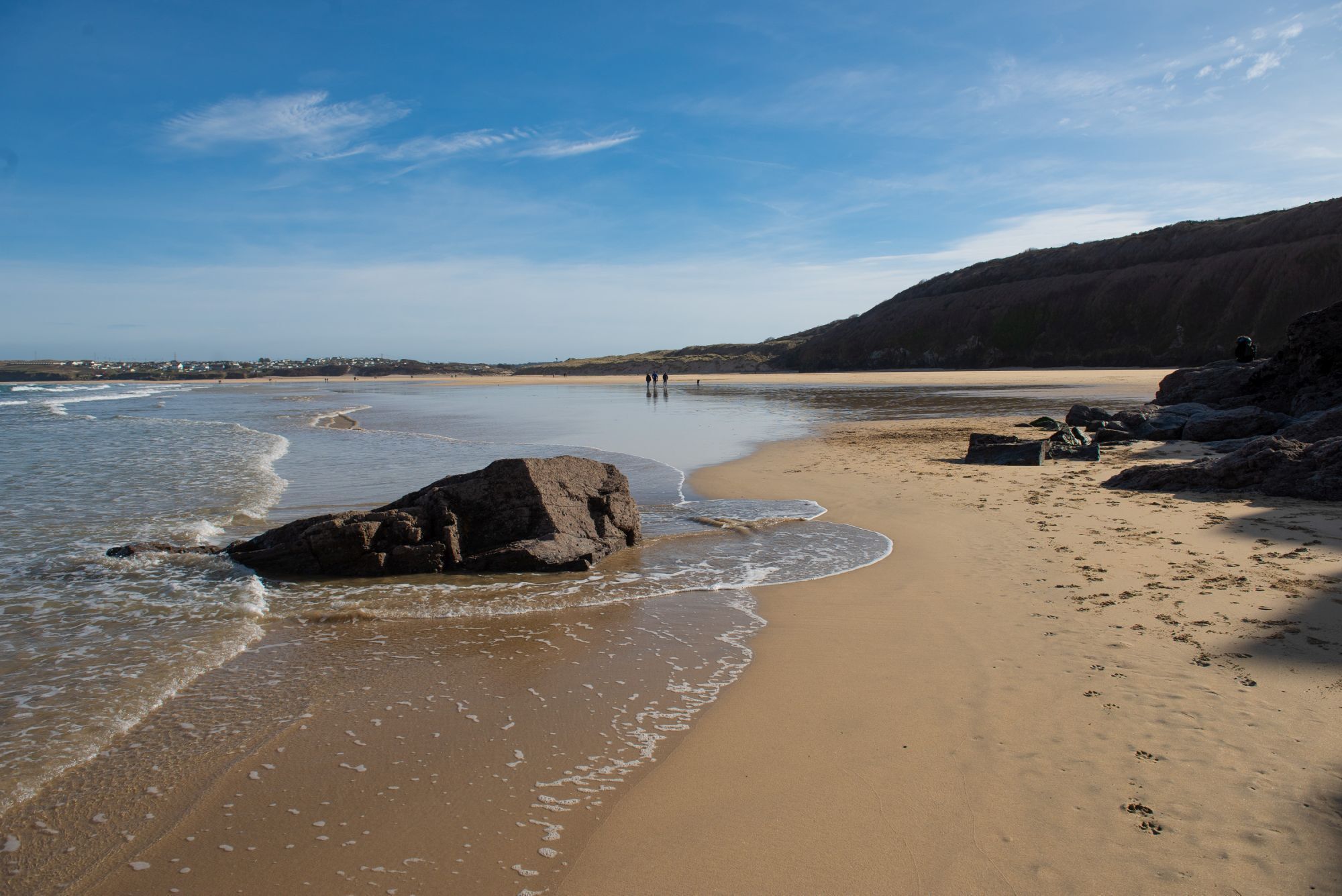 A beach with clear skies, a large rock in the foreground, and a sandy shore.