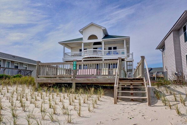 Ein Strandhaus mit Balkon, einer Holzterrasse und einem sandigen Strand im Vordergrund.