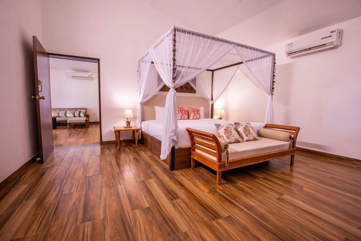 A bedroom with a four-poster bed, wooden flooring, a bedside table with a lamp, and an air conditioning unit on the wall.