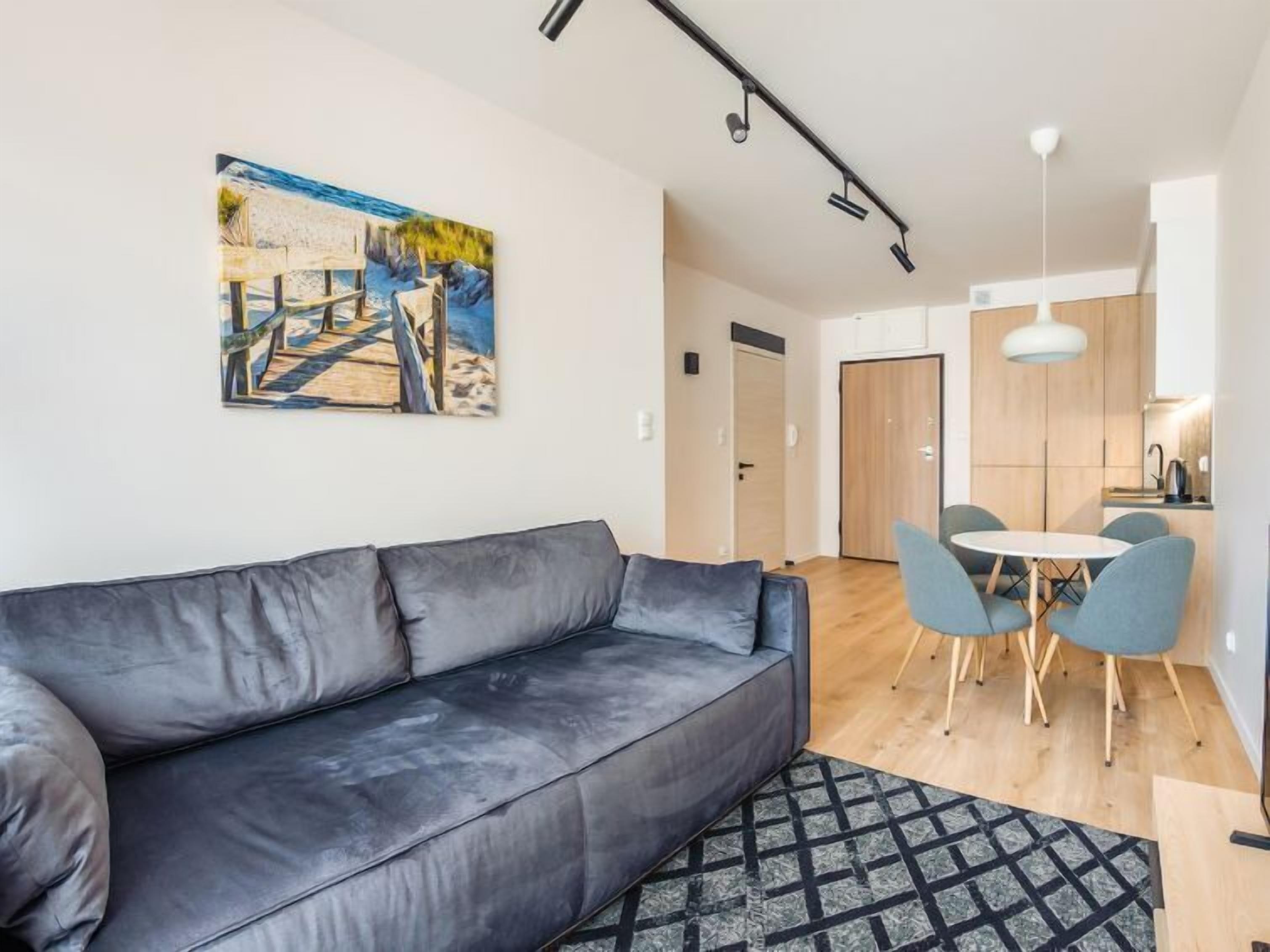 Un salón moderno con un sofá gris, un cuadro de una escena de playa, una zona de comedor con una mesa redonda y sillas azules, y una cocina al fondo.