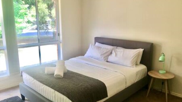 A neatly made bed with white and gray linens, a small bedside table with a green lamp, and a window with a view of greenery outside. - Spacious townhouse located in the CBD! (East Wagga Wagga)
