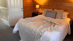 Une chambre confortable avec un plafond en bois, une porte blanche, une table de chevet avec une lampe et un lit recouvert d’une couette blanche et agrémenté d’oreillers.
