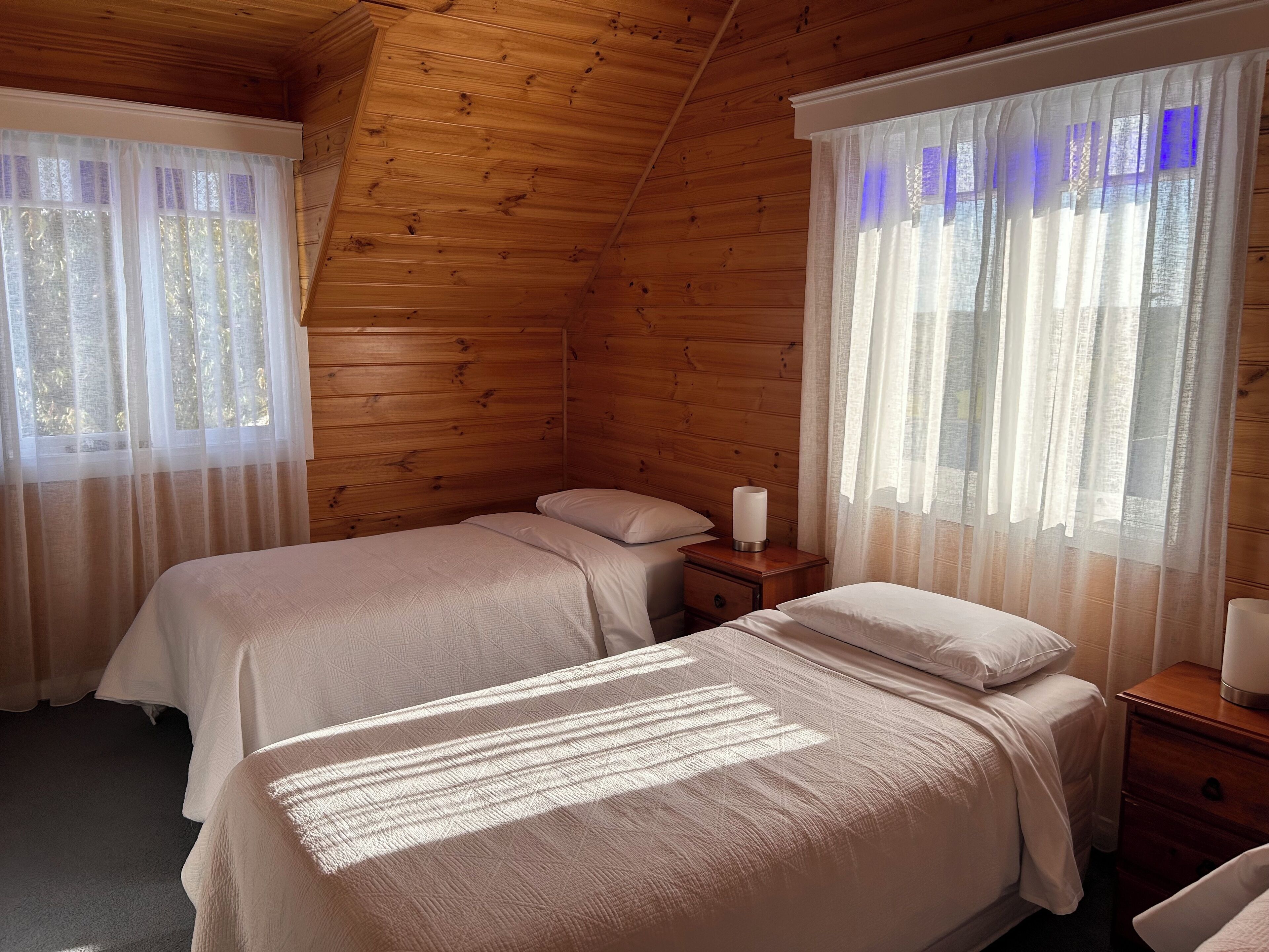 A cozy room with wooden walls, two beds with white linens, and a nightstand with a lamp.