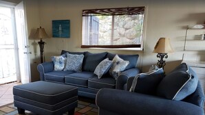 A living room with a sofa, ottoman, and armchair, a window with a view, and a painting on the wall.