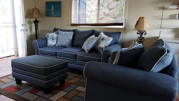 Relax in this inviting living space with a sofa set and ottoman on a patterned rug, offering a view of lush greenery and a beautiful painting on the wall.