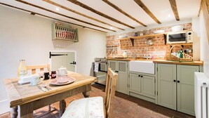 A kitchen with a wooden table, a chair, a brick wall, a microwave, and a sink. - Oast Barn Cottage by Bloom Stays -  2 guests  in 1 bedroom (Faversham)