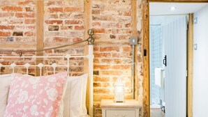 A bedroom with a brick wall, a white bed with floral pillows, a bedside table with a lantern, and a wooden door leading to a bathroom. - Oast Barn Cottage by Bloom Stays -  2 guests  in 1 bedroom (Faversham)