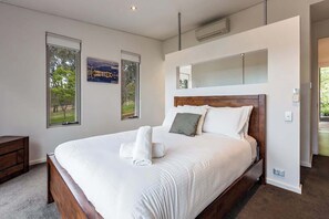 A modern bedroom with a large bed, a wooden headboard, and a view of a park through a window.