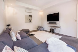 A modern living room with a grey sofa, a flat-screen TV mounted on the wall, a white rug, and a small black side table.