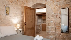 Une chambre avec un mur en pierre, une porte en bois, un lit recouvert de draps blancs, une table de chevet avec une lampe et un miroir accroché au mur.