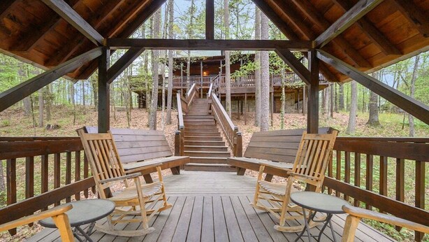 A wooden deck with rocking chairs and a staircase leading to a cabin in a forest.