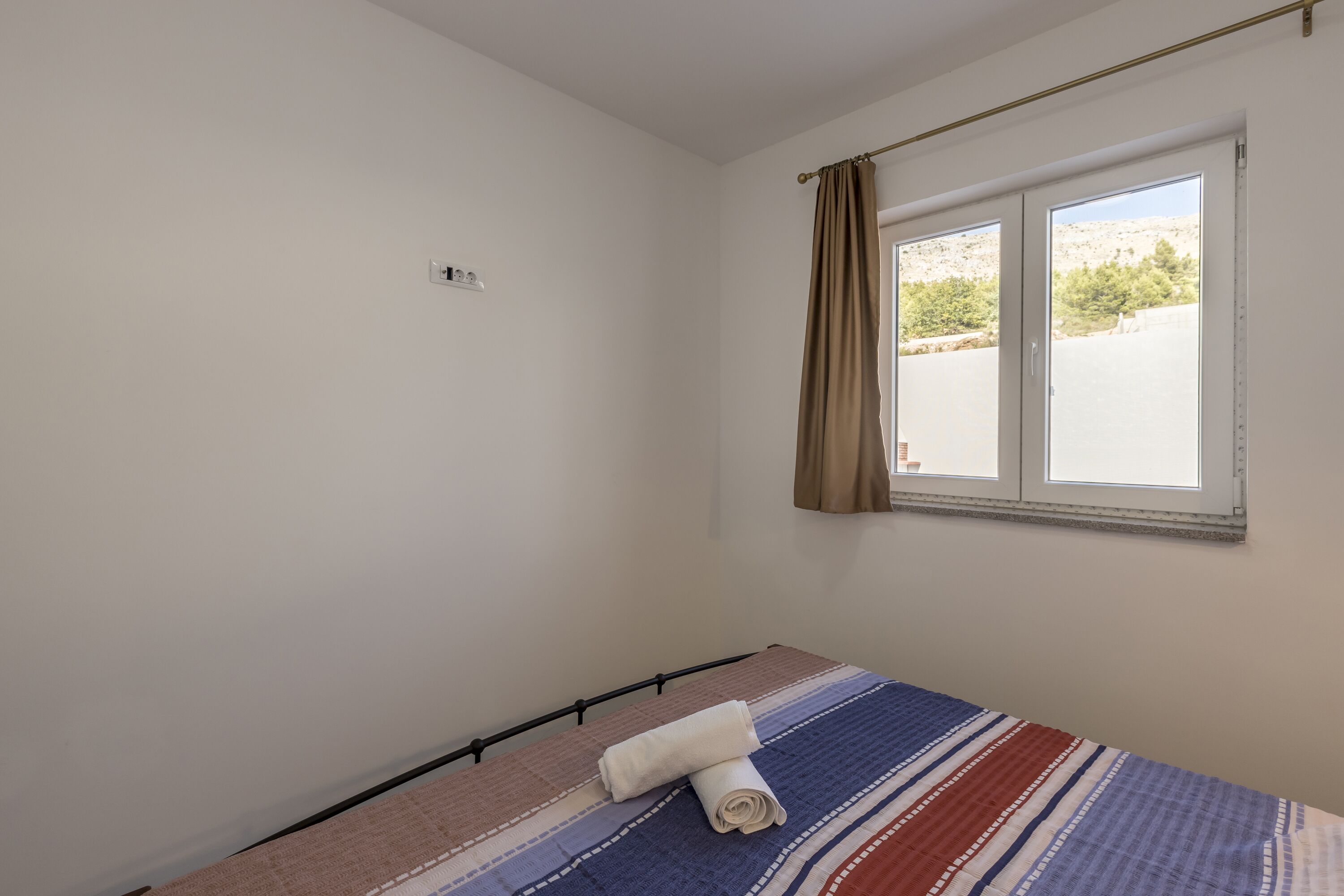 A single bed with a striped bedspread and a rolled towel, a window with curtains, and a white wall with a light switch.