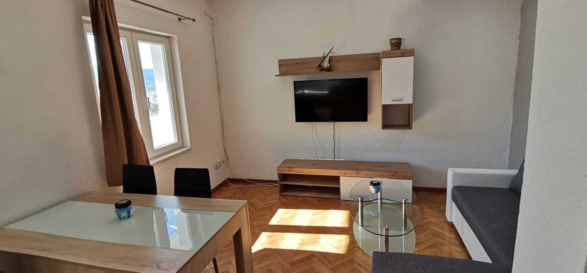 A modern living room with a flat-screen TV mounted on the wall, a wooden coffee table, a glass-top dining table, and a grey sofa.