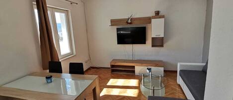 A modern living room with a flat-screen TV mounted on the wall, a wooden coffee table, a glass-top dining table, and a grey sofa.