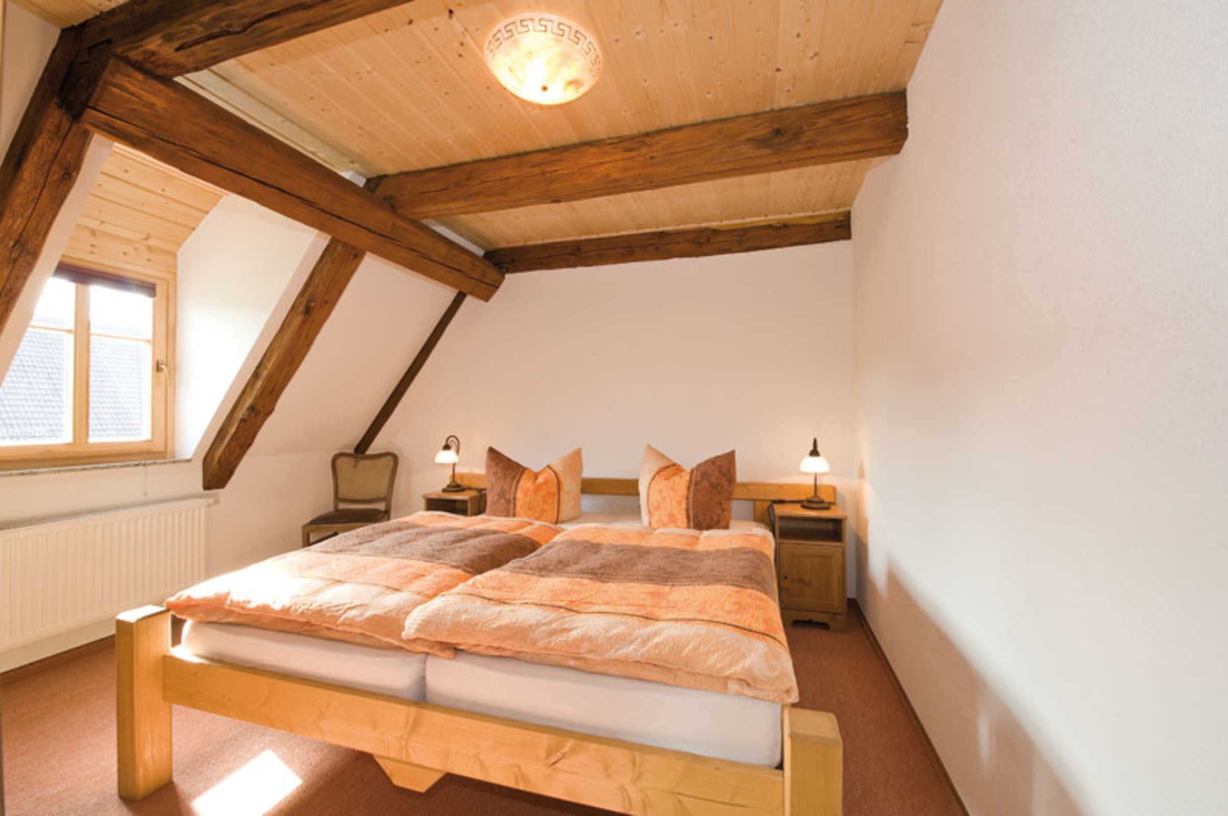 Un dormitorio con cama de madera, dos mesitas de noche con lámparas, una ventana con vistas y un techo inclinado con vigas a la vista.