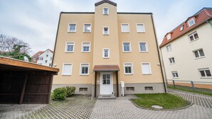 Ein mehrgeschossiges Gebäude mit zentralem Eingang, einer kleinen Veranda und einem Ziegelweg, der zum Eingang führt.