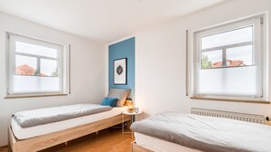 Ein Zimmer mit zwei Holzbetten, einem kleinen Tisch mit Lampe, einem gerahmten Bild an der Wand und großen Fenstern mit Jalousien.