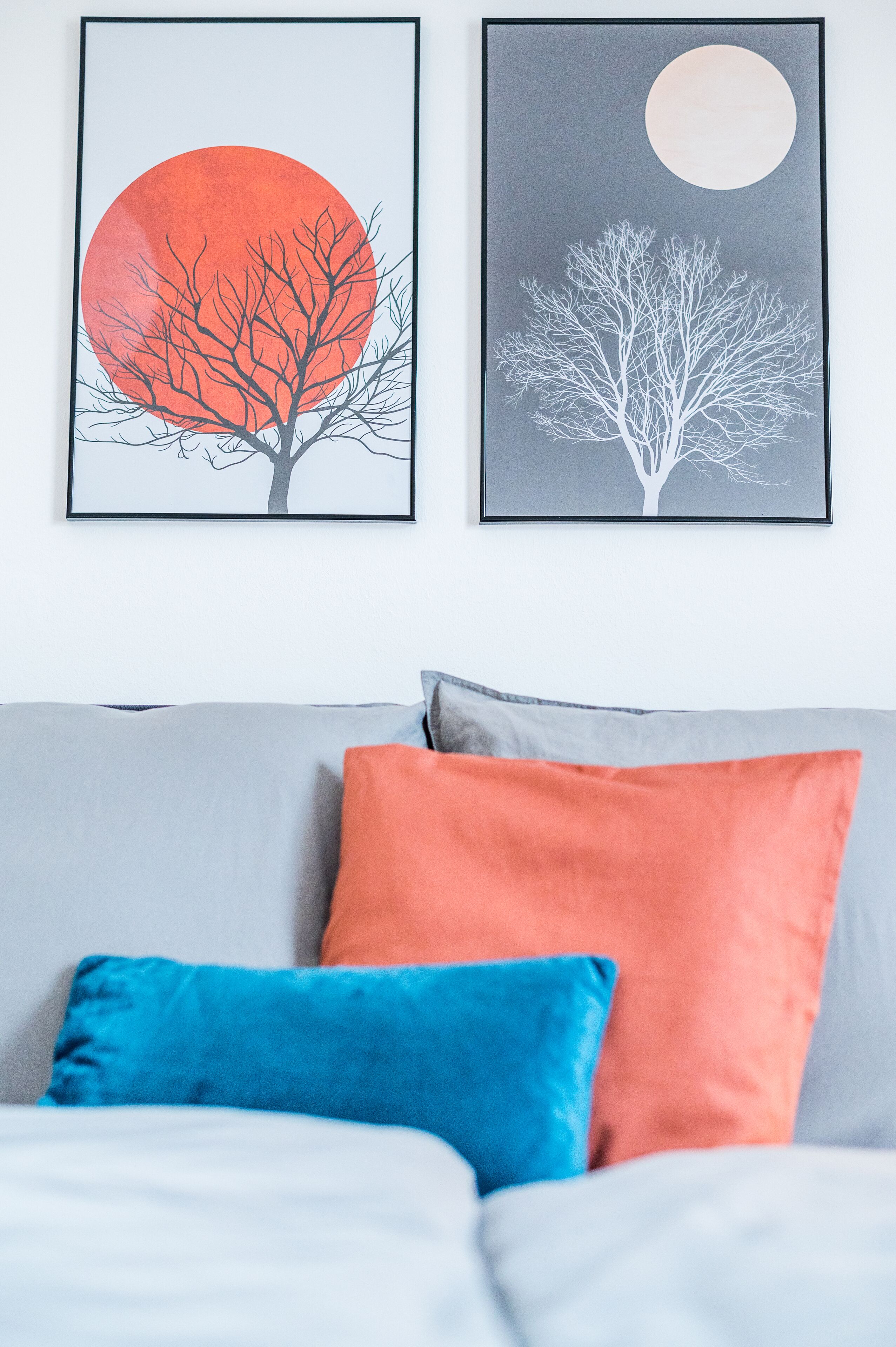 A bedroom with two framed artworks depicting trees and moons, a grey sofa, and two pillows in blue and orange.