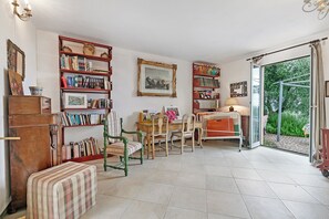 A cozy living room with a bookshelf, a dining table with chairs, a wooden cabinet, and a large window with a view of greenery.