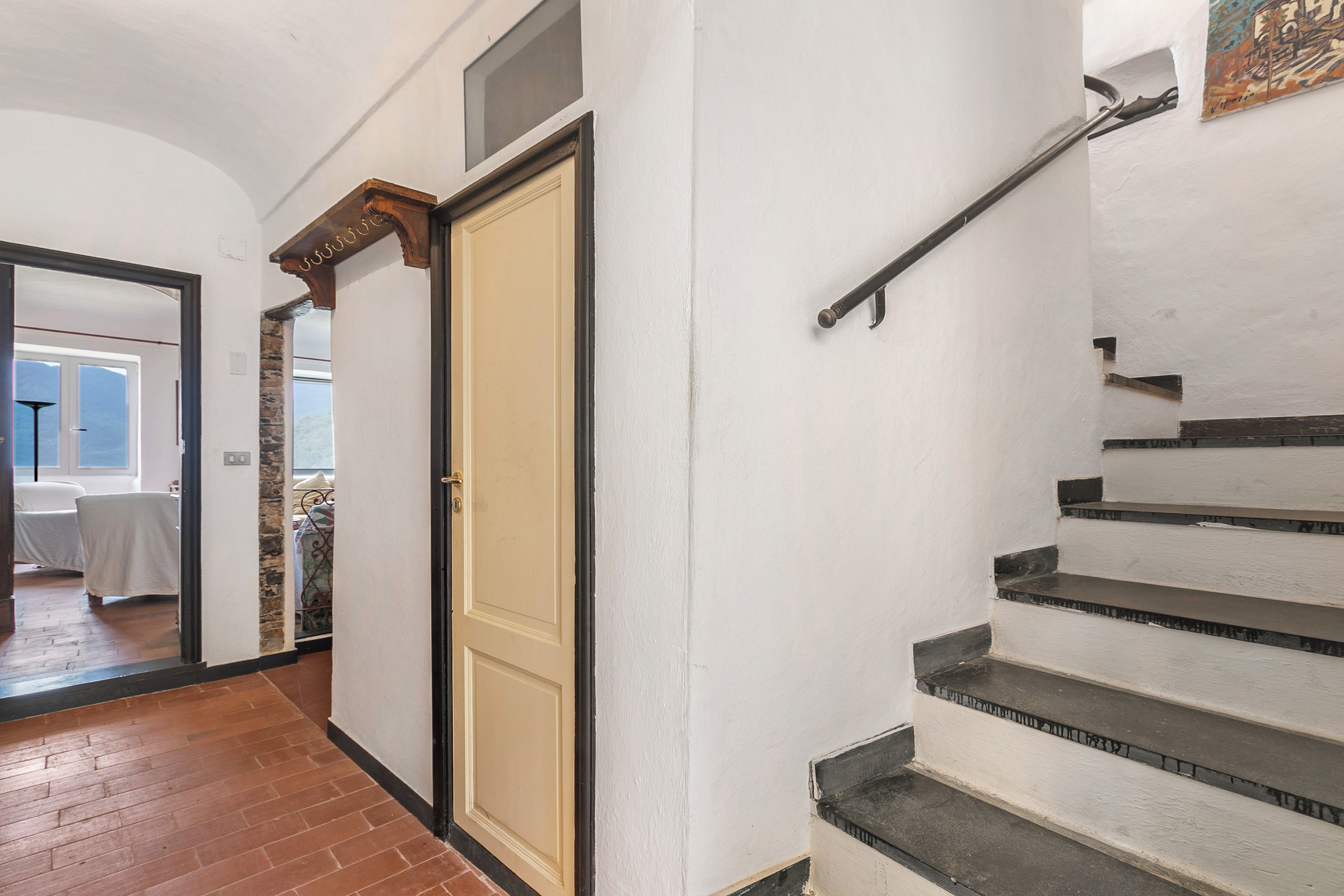 A staircase with a handrail, a door with a wooden frame, and a view into a room with a bed and a window.