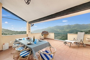 Un balcone con un tavolo apparecchiato per mangiare, sedie, un barbecue e una vista panoramica sulle montagne.