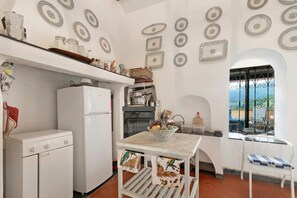 A compact kitchen with a white refrigerator, stove, and oven. A dining table with a bowl of fruit and a chair is visible. Decorative plates hang on the wall, and a window with a wrought iron gate is present. - Ca Veggia for Nature Lovers With Views & Garden (Stellanello)