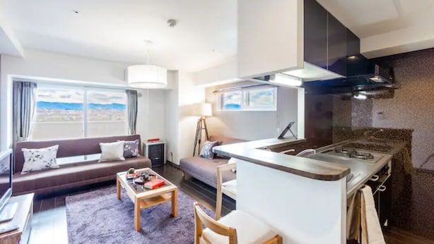 A modern living room with a sofa, a coffee table, and a dining area. There is a kitchen with a stove and a view of the city. - Diva (Sapporo)